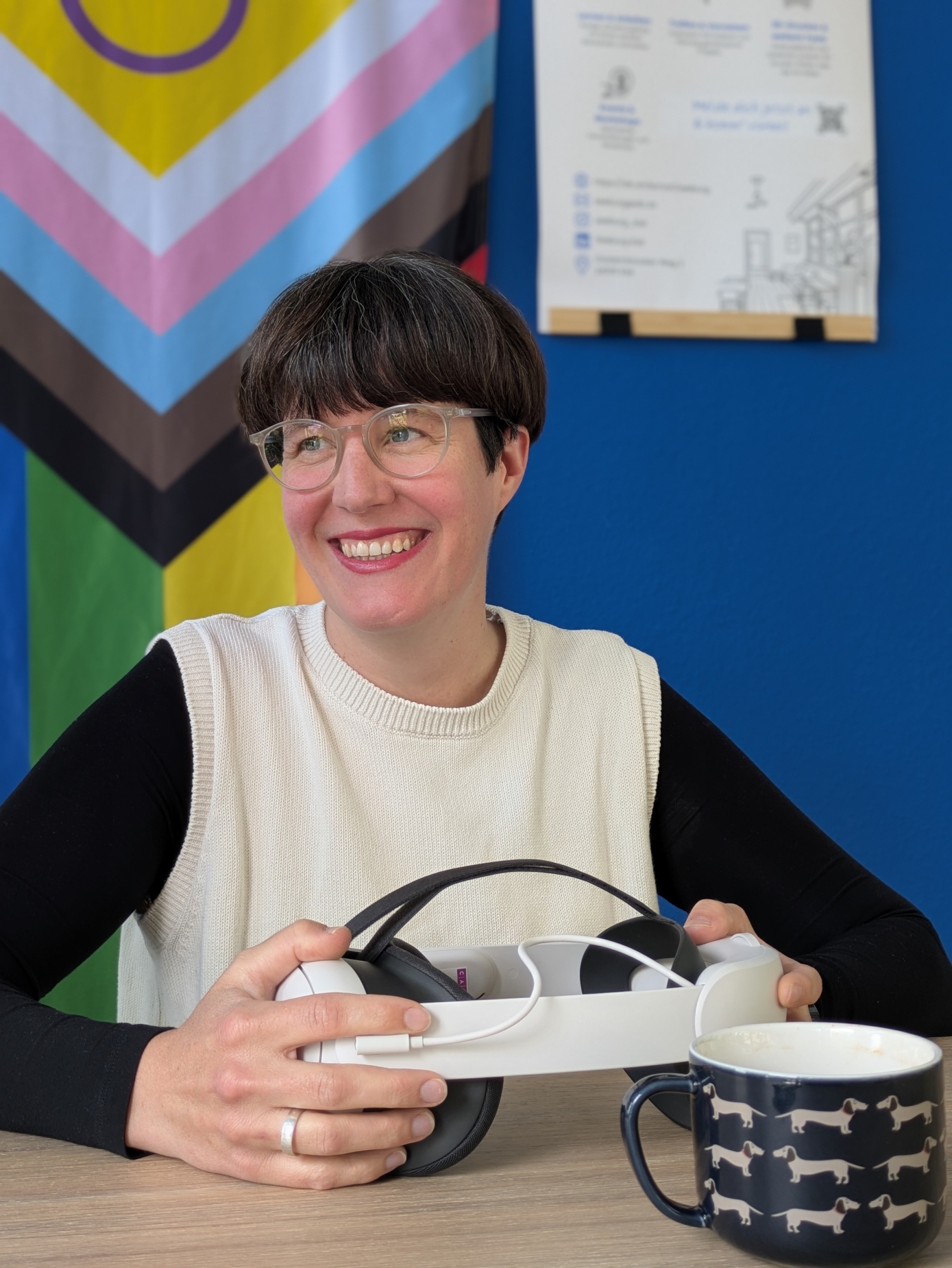 Eine Frau mit kurzen dunklen Haaren, heller Brille sitzt mit einer VR Brille an einem Tisch.